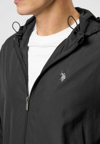 Mees, kes kannab musta kapuutsiga lukuga jope ja all paistab valge särk, kus on rinnal väike polo mängija logo.