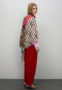 Bufanda de estampado de leopardo y rayas con toques rojos, que presenta una textura suave. Llevada sobre una camiseta de manga larga rosa y pantalones rojos.