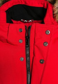 Rød parka med en hætte beklædt med falsk pels, prydet med en sort lynlås og skinnende sølvtryklåse på et tekstureret stof.