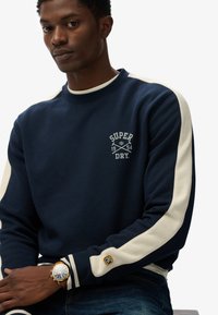 Sudadera de color navy con franjas en crema en los hombros, que presenta el logo "SUPER DRY" y el año. Tejido suave con puños acanalados. Lleva un reloj.