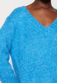 Persona che indossa un maglione V-collo in maglia pelosa di un brillante blu, che mostra l'area del petto e del braccio superiore su uno sfondo neutro.