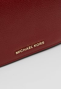 Borsa a mano in pelle rossa con superficie texture; presenta un logo dorato "MICHAEL KORS" sul fronte, evidenziando la sua identità di marca.