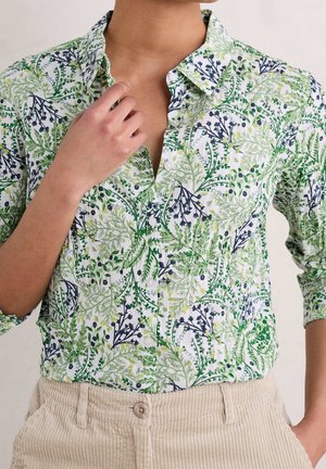 Nainen, jolla on valkoinen napillinen paita, jossa vihreä- ja mustalehtikuvio, yhdistettynä beigen värisiin puuvillahousuihin, käsi koskettaa kaulusta.