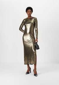 Metalická zlatá dlouhá šaty s přiléhavým střihem a bočním rozparkem, doplněné černými podpatky a malou černou kabelkou.