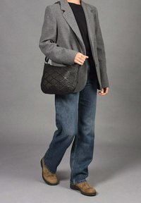 Bolso de hombro negro tejido con patrón de diamantes, llevado por una persona con un blazer gris, top negro, jeans azules y botas marrones.
