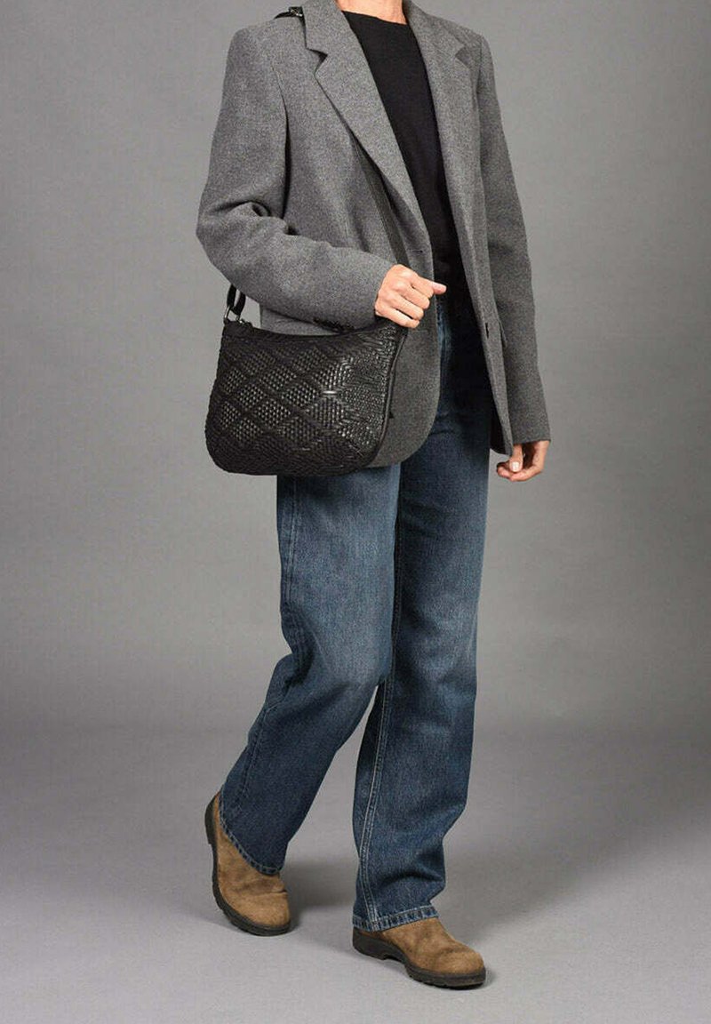 Bolso de hombro negro tejido con patrón de diamantes, llevado por una persona con un blazer gris, top negro, jeans azules y botas marrones.