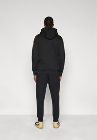 Μαύρη φούτερ μπλούζα με κουκούλα και λογότυπα, συνοδευόμενη από αντίστοιχο παντελόνι φόρμας. Η εμφάνιση περιλαμβάνει ραβδωτούς μανσέτες και casual παπούτσια.