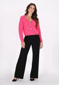 Cardigan rosa con motivo a rombi e bottoni decorativi, abbinato a pantaloni neri a gamba larga e tacchi neri. Tessuto liscio e design aderente.