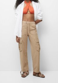 Pantalons cargo beiges avec poches latérales et taille élastique, associés à un haut de bikini orange et une chemise en broderie blanche, portant des sandales marron.