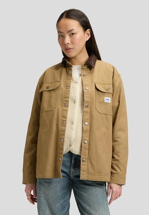 Junge Frau trägt eine hellbraune Jacke mit Knöpfen und Klappentaschen sowie eine Lammfellweste darunter, kombiniert mit blauen Jeans.