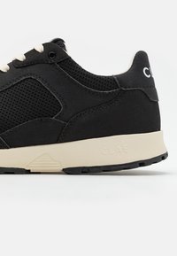 Svart sneaker med mesh-overdel og lærdetaljer, kremfarget mellomsåle, strukturert overflate og en logo på hælen.