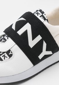 Weiße Sneaker mit strukturiertem Lederfinish, ausgestattet mit einem schwarzen elastischen Band mit weißen Buchstaben und einem gemusterten Fersenanhänger. Gummisohle.