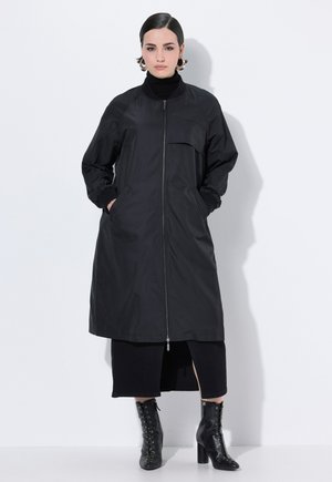 Manteau long noir avec une fermeture éclair, des poignets côtelés et des poches latérales. Tissu texturé avec un design minimaliste et une seule poche avant.