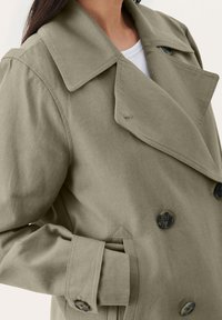 Vihreä oliivinvihreä kaksirivinen trench coat, jossa on teksturoitu kangas, suuria nappeja, leveät kaulukset ja rento istuvuus, jossa on hihansuut.