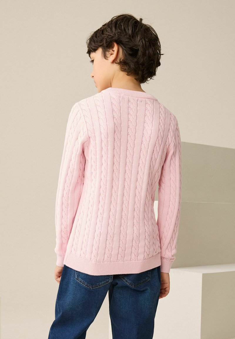 Lyserød kabelstrikket sweater med rund halsudskæring, lange ærmer og ribbede manchetter. Brugt sammen med mørkeblå jeans, der viser et tekstureret design.