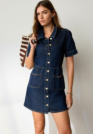 Junge Frau trägt ein ärmelloses Jeans-Knopfkleid und hält eine gestreifte Tragetasche über der Schulter vor einfarbigem Hintergrund.