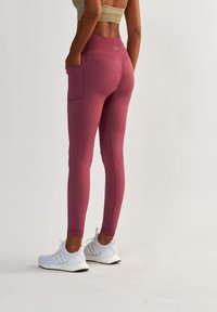 Leggings taille haute, ajustés, de couleur bordeaux avec des poches latérales. Associés à des chaussures de sport blanches, mettant en valeur une texture de tissu lisse.