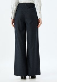 Donkerblauwe wijde broek met een gestructureerde stof, voorzien van een gladde tailleband en twee achterzakken. Draagt een paar zwarte hoge hakken.