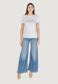 Maglietta bianca con maniche corte e logo grigio "GUESS", abbinata a jeans a zampa d'elefante di colore azzurro chiaro e tacchi nudi su uno sfondo neutro.