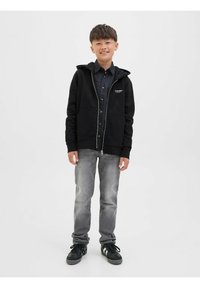 Crna zip-up hoodie s kapuljačom, prednjim džepovima i detaljem logotipa, u kombinaciji s sivim trapericama i crnim tenisicama. Glatka tekstura, ležeran dizajn.