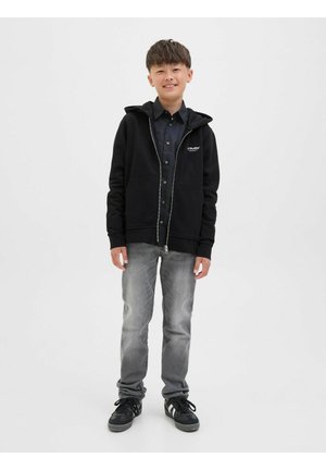 Haut à zip noir avec capuche, poches avant et détail de logo, associé à un jean gris et des baskets noires. Texture lisse, design décontracté.