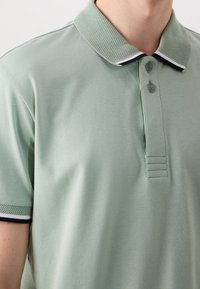 Homme portant un polo vert clair avec un col texturé et des bords de manches ornés de rayures noires et blanches, deux boutons fermés.