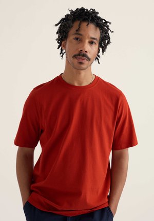 Mężczyzna z kręconymi włosami i wąsami, ubrany w prostą czerwoną koszulkę z krótkim rękawem, stoi z rękami w kieszeniach na jasnym tle.