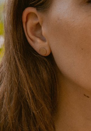 Goldene runde Steckerohrringe, die im Ohr einer Frau getragen werden. Die Oberfläche ist glatt und sie sind klein, was den natürlichen Hautton unterstreicht.