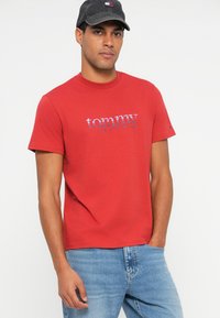 Camiseta de algodón roja con cuello redondo y mangas cortas. Presenta el logo "tommy JEANS" en blanco y azul en el pecho.
