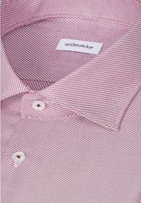 Gros plan sur le col d'une chemise texturée rose avec des boutons blancs et une étiquette de marque "seidensticker" visible.