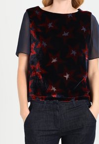 Top de manga corta en terciopelo azul marino con estampados de estrellas rojas y grises, con un corte relajado y mangas transparentes, combinado con jeans oscuros.
