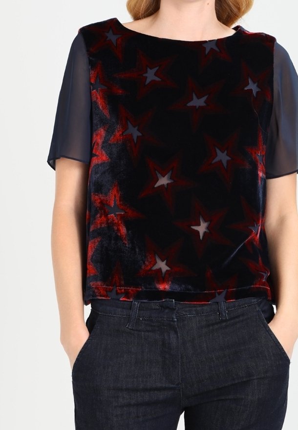 Top de manga corta en terciopelo azul marino con estampados de estrellas rojas y grises, con un corte relajado y mangas transparentes, combinado con jeans oscuros.