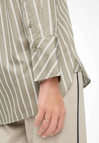 Camicia a righe verde oliva con linee verticali bianche, caratterizzata da un polsino con bottoni e una vestibilità comoda. Il tessuto appare liscio e leggero.