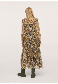 Nainen, jolla on pitkät vaaleat hiukset ja yllään pitkä, läpikuultava kukkakuosisella röyhelömekko, yhdistettynä tumma vihreisiin, järeisiin saappaisiin, seisoo vaalealla taustalla.