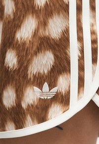 Καφέ και λευκό υφάσμα με λευκό λογότυπο Adidas και τρεις λευκές κάθετες ρίγες περιβαλλόμενες από λευκό περίγραμμα.
