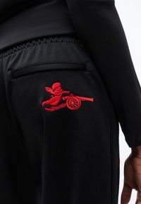 Pantalon noir avec taille élastique et un chérubin rouge brodé, visant un canon, sur la cuisse supérieure près d'une poche passepoilée.