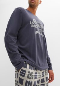T-shirt a maniche lunghe di colore blu navy con scollatura a V e stampa grafica argentata, abbinato a pantaloni grigi chiari con un motivo a griglia scura e linee orizzontali.