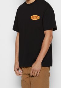 Muž v černém tričku s malým žlutým a červeným logem Carhartt WIP na hrudi a béžových prošívaných kalhotách.