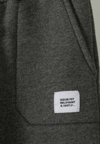 Graue Stoff-Jogginghosen mit strukturiertem Oberflächen, Seitentasche und einem weißen eingenähten Etikett, das "ISSUE/07 RELEVANT S.1997//" liest.
