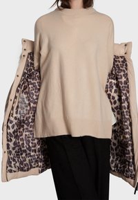 Beige stickad tröja med hög hals, bärs under en ljusjacka med leopardmönstrad foder.
