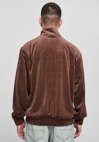 Brun fløyelsjakke med høyt krage, elastisk midje og lange ermer; glatt tekstur og avslappet passform. Brukt over lyseblå jeans.