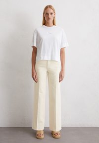 Weißes kurzärmeliges Cropped-T-Shirt mit kleinem schwarzem Logo. Kombiniert mit cremefarbenen weit geschnittenen Hosen und beigefarbenen Plateausandalen.