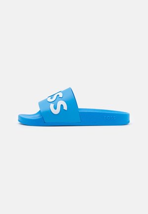 Toboggan en caoutchouc bleu avec une large sangle arborant un logo blanc en relief. Semelle intérieure texturée et semelle lisse. Design minimaliste avec un matériau durable.