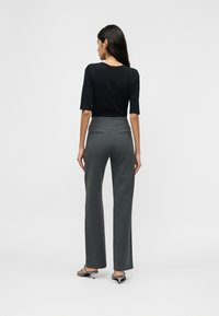 Γυναίκα με μακριά σκούρα μαλλιά, που στέκεται γυρισμένη προς τα πίσω, φοράει μαύρο τοπ με μανίκια έως τον αγκώνα, γκρι παντελόνι καλοραμμένο και μαύρα σανδάλια με τακούνι.