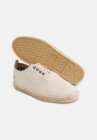 Espadrilles de lienzo beige con suelas de yute trenzado, cordones blancos y un logo azul en el lateral. La suela exterior presenta un patrón geométrico.