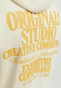 Sudadera crema con un gran diseño de texto amarillo. El texto incluye "ORIGINALS," "STUDIO," y "CREATIVE COMMUNITY." Tela suave con capucha.