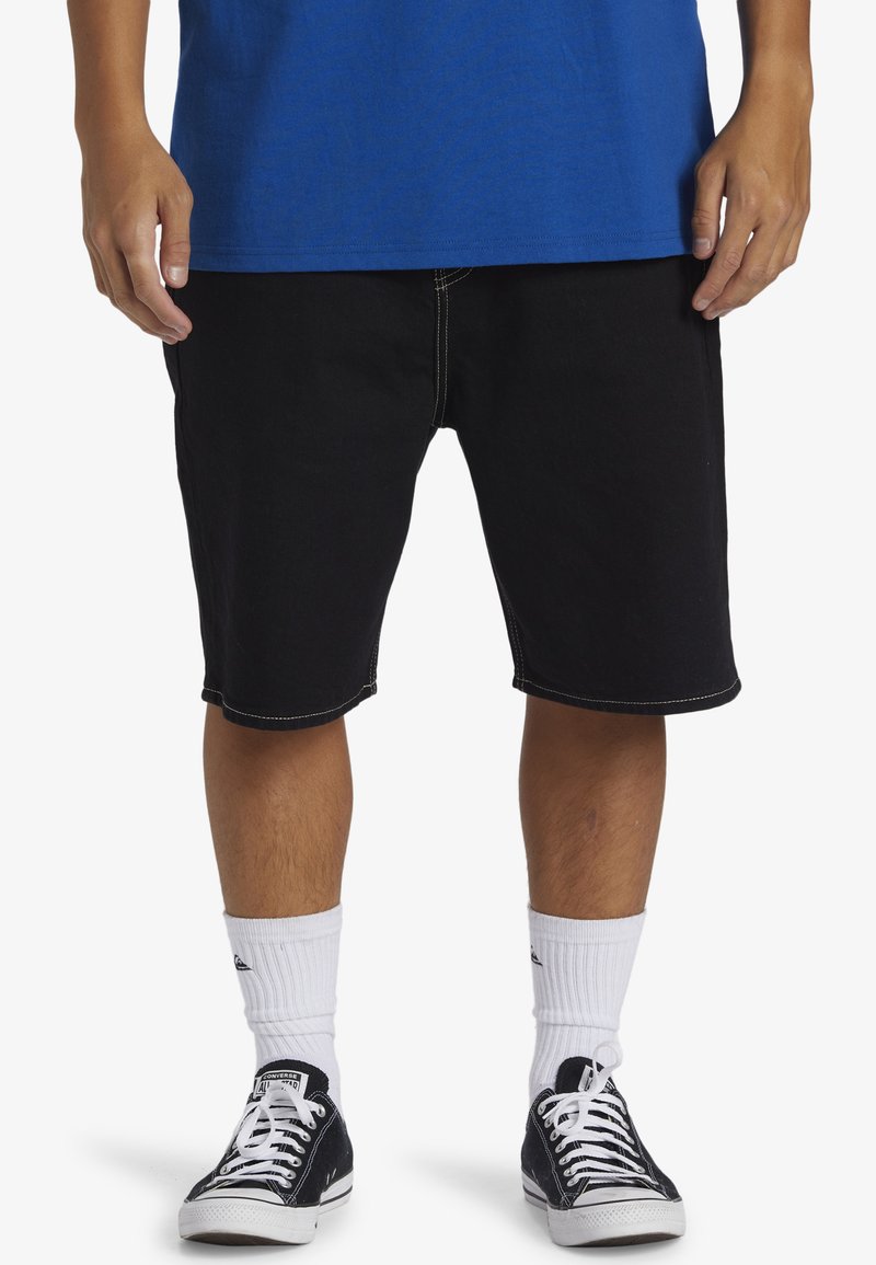 Shorts de mezclilla negra con costuras en contraste, de longitud a la rodilla. Combinados con calcetines blancos hasta el crew y zapatillas negras de caña baja. Camiseta azul visible en la parte superior.