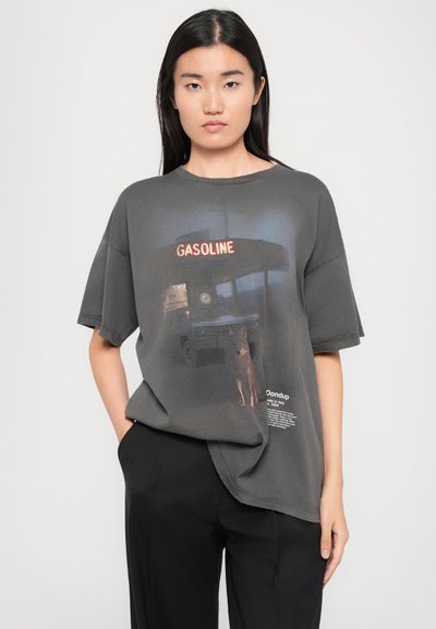 Gray oversized t-shirt with a graphic print of a gas station and dog. Features wide sleeves, raw hem, and text detailing at the bottom.