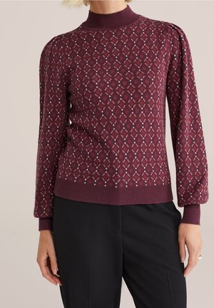 Vrouw draagt een bordeauxrode trui met lange mouwen en hoogsluitende hals, met patroon, en zwarte broek, staand tegen een effen achtergrond.