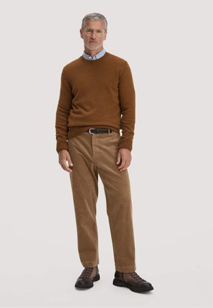 Mann mit grauen Haaren und Brille, der einen braunen Pullover, ein blaues Hemd mit Kragen, beige Cordhosen, schwarze Stiefel und einen geflochtenen Gürtel trägt, steht vor weißem Hintergrund.